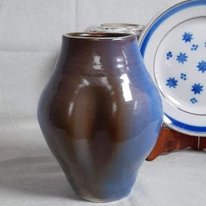 May include: A handmade ceramic vase with a distinctive form and a gradient colour scheme. The vase fades from a deep brown at the top to a soft blue at the base. A decorative plate with blue star patterns is in the background.