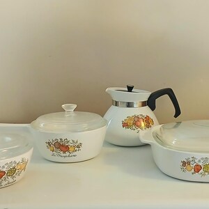 May include: A set of four white ceramic cookware pieces with floral designs and glass lids. The set includes a teapot, a casserole dish, and two smaller pots. The cookware features a floral pattern with red, yellow, and green accents.