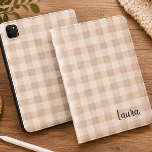 Peut inclure: Deux étuis pour tablette avec un motif à carreaux vichy beige et blanc. Un étui présente une découpe pour l'appareil photo, tandis que l'autre est de style folio avec le prénom Laura imprimé en écriture noire en bas à droite.