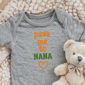 May include: A gray baby onesie with the text "pass me to NANA" in orange and green letters with a heart below. The onesie is lying on a white fuzzy blanket with a teddy bear.