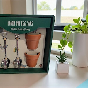 May include: A green box set with the text "PLANT POT EGG CUPS" and "With 2 shovel spoons". Inside are two small terracotta-coloured pots and two silver shovel-shaped spoons. The background includes a window and two small potted plants.