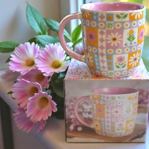 May include: A patterned ceramic mug with a pink handle and interior. The mug features a patchwork design with daisies and other flowers in pink, yellow, and blue. The mug sits on a matching box. Pink flowers are arranged nearby.