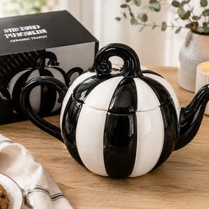 May include: A ceramic teapot shaped like a pumpkin with alternating vertical black and white glossy stripes. The teapot features a curved black handle, a matching black spout, and a lid with a black stem-shaped handle. The box is labelled STRIPED PUMPKIN.