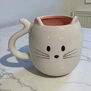 May include: White ceramic cat-shaped mug with a pink interior. The mug features black eyes, a nose, and whiskers. The handle is shaped like a cat's tail. A charming and whimsical design for cat lovers.