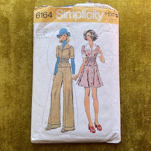 Puede incluir: Un sobre de patrón de costura Simplicity, número 6164, con ilustraciones de dos conjuntos: un mono color canela con cuello alto azul y sombrero, y un vestido a rayas rojas y blancas con cuello blanco. El texto incluye talla 13/14, busto 33.5 y cintura 26.