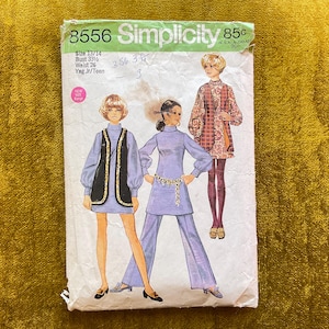 Op de afbeelding: Een vintage Simplicity naaipatroon envelop, nummer 8556, met illustraties van drie outfits waaronder een vest, een tuniek en een broek. De witte papieren envelop toont de tekst Simplicity 8556, maat 13/14, buste 33,5 en taille 26.