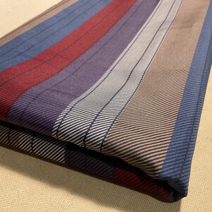 May include: Folded fabric with vertical stripes in shades of blue, red, purple, white, and brown. The stripes have a subtle diagonal texture. The fabric is folded over, showing the pattern on the top layer.