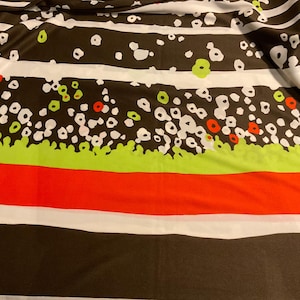May include: Fabric with a repeating pattern of white, green, and red shapes on a dark brown background. The design includes white dots, green and red flower-like shapes, and horizontal stripes.