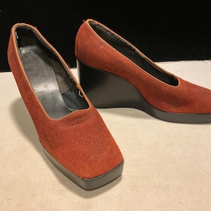 May include: A pair of rust-colored suede wedge heels. The shoes have a square toe and a black wedge heel. The interior of the shoe is black and the sole is black. The shoes are set against a neutral background.