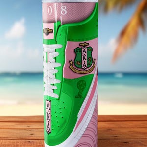 May include: A tall, insulated tumbler featuring a green and pink sneaker design with white laces. The tumbler includes the letters "AKA" and a crest, with a straw. The background shows a beach scene.
