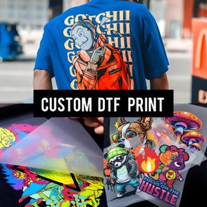 May include: A blue t-shirt with a graphic of a person in an orange jacket and a mask. The shirt has the text "GOCHII" and "CUSTOM DTF PRINT". Below are several colorful graphic prints, including a panda, dog, and mushrooms.