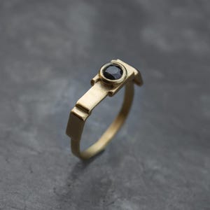May include: A gold-colored ring with a black gemstone. The ring has a unique geometric design, featuring a rectangular band and a circular setting for the dark stone. The ring's design is modern and minimalist.