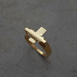 May include: A gold-colored ring with a geometric design. The ring features a rectangular bar with two perpendicular rectangular accents. The ring is set against a gray background.