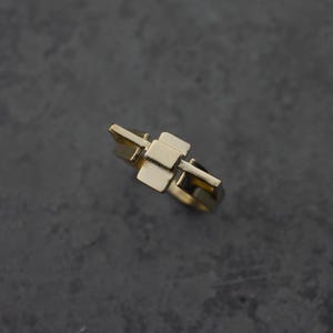 May include: A gold-colored ring with a geometric design. The ring features a series of rectangular shapes connected to the band. The ring is set against a dark gray background. The ring is a piece of jewelry.
