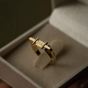 May include: A gold-colored ring with a geometric design. The ring features a rectangular center section with two perpendicular bars on either side. The ring is displayed in a light-colored box.