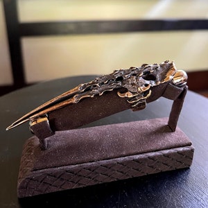 Puede incluir: Un anillo detallado, texturizado, de color bronce en forma de garra, exhibido en un soporte de terciopelo marrón.