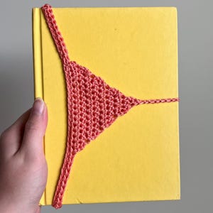 May include: A bright yellow hardcover book secured with a coral-colored, triangular crochet bookmark. The bookmark features a textured, hand-stitched pattern that wraps around the cover, providing a functional way to keep the book closed.