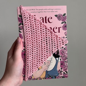 May include: A triangular light pink crochet bookmark made of yarn with a textured stitch pattern. The bookmark is placed over the corner of a book cover featuring a floral design with pink roses and an illustration of two people.