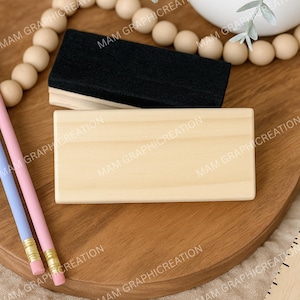 May include: A light-colored wooden rectangular sign and a black felt eraser sit on a wooden surface. Two pencils, one pink and one blue, are positioned on the left side. The background includes a wooden bead garland.