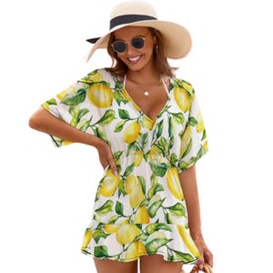 May include: A white, short-sleeved dress with a V-neck and a lemon print. The dress has a ruffled hem and a drawstring waist. The person in the image is wearing a straw hat with a black band and sunglasses.