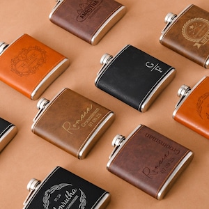 May include: An assortment of personalized flasks in various colors, including brown, black, and orange. Each flask has a silver cap and trim, with custom engravings such as names, dates, and decorative designs. The flasks are arranged on a neutral background.