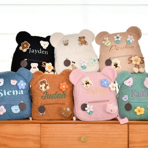 May include: A collection of teddy bear-shaped backpacks in various colors, including black, white, blue, brown, pink, and green. Each backpack features a name embroidered on the front, along with decorative floral and bear appliqués. The backpacks have a zippered front pocket.