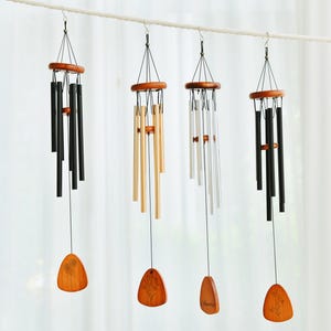 May include: Four wind chimes hanging from a rope. Each chime has a wooden top and a wooden wind catcher. The chimes are black, gold, and silver. The wind catchers have a tree design.