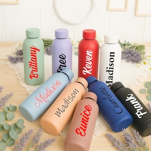 Puede incluir: Una colección de botellas de agua aisladas en varios colores, incluyendo verde, morado, rojo, blanco, azul, melocotón y marrón. Cada botella tiene un nombre diferente impreso en un color contrastante. Las botellas están dispuestas sobre una superficie de madera.