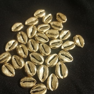 May include: A collection of gold-colored cowrie shells, each with a distinctive oval shape and a central slit. The shells are scattered on a dark surface, creating a contrast that highlights their metallic sheen. These shells are often used in jewelry and decorative crafts.