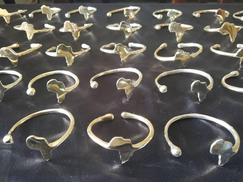 May include: A collection of open cuff bracelets with a map of Africa as the centerpiece. The bracelets are made of a shiny, gold-colored metal and are arranged on a dark surface. The bracelets have a simple, elegant design.