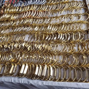 Op de afbeelding: Een grote collectie goudkleurige metalen armbanden gerangschikt in nette, parallelle rijen. Elke armband heeft een gladde, gepolijste afwerking met een consistente ronde vorm, wat zorgt voor een dichte, uniforme weergave van metalen sieraden.