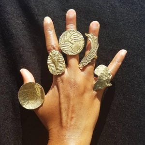 May include: A hand displaying several gold-colored rings. The rings feature various designs, including a pharaoh, a textured circle, a wing, and an African continent silhouette. The rings are of different sizes and shapes.