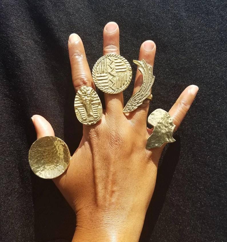 May include: A hand displaying several gold-colored rings. The rings feature various designs, including a pharaoh, a textured circle, a wing, and an African continent silhouette. The rings are of different sizes and shapes.