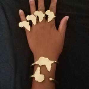 May include: Gold-colored jewelry set featuring rings and bracelets. The rings are shaped like the African continent and are worn on the fingers. The bracelets also feature the continent's shape. The jewelry is displayed on a dark background.