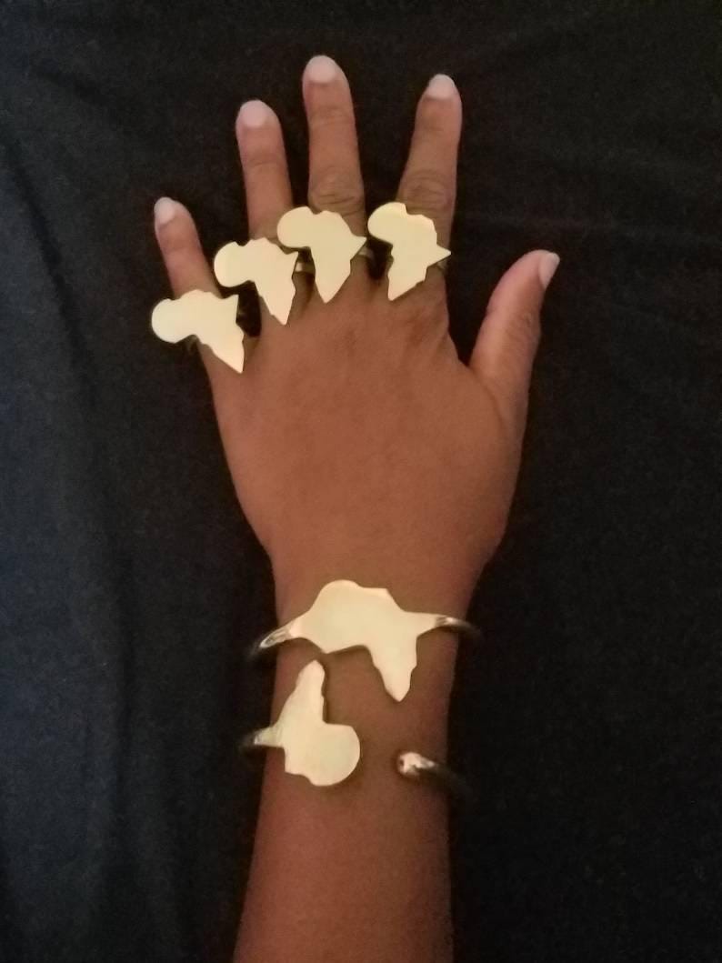 May include: Gold-colored jewelry set featuring rings and bracelets. The rings are shaped like the African continent and are worn on the fingers. The bracelets also feature the continent's shape. The jewelry is displayed on a dark background.