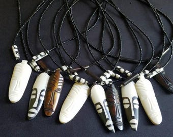 May include: A collection of handcrafted necklaces featuring carved mask pendants. The pendants are in various colors, including white, brown, and black, strung on black beaded cords. The masks have detailed facial features and are of varying sizes.