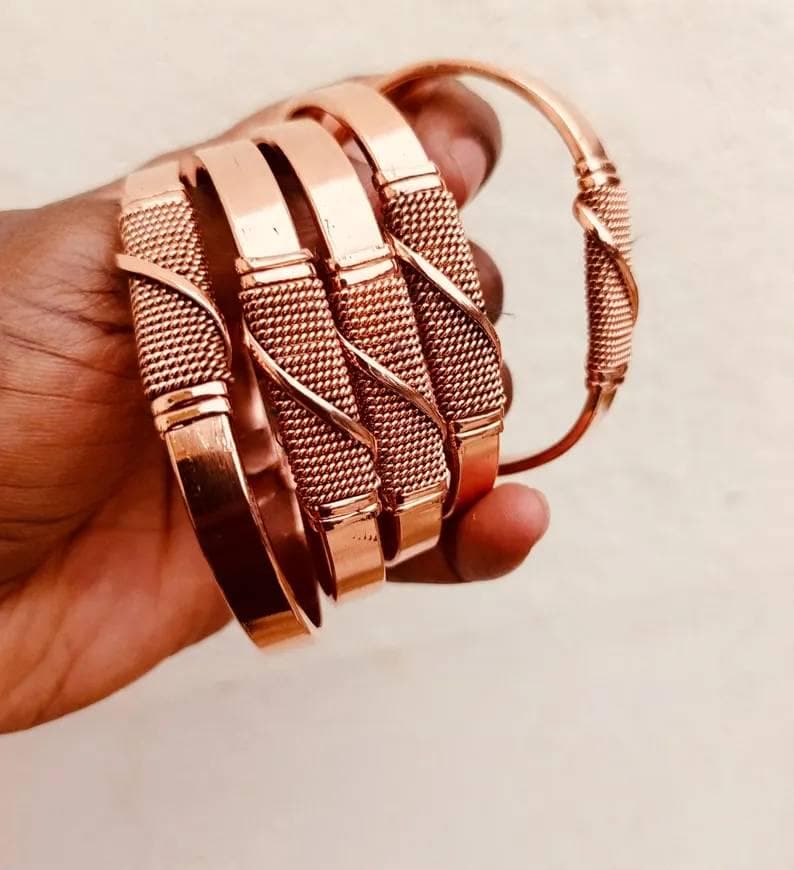 May include: A collection of copper-colored bangles. The bracelets have a mix of smooth, flat bands and sections with a woven texture. One bangle is fully visible, while the others are partially visible, held in a hand.