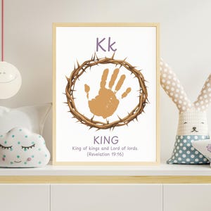May include: A framed print with the letter "Kk" in purple, a handprint within a crown of thorns, and the word "KING" below. The text "King of kings and Lord of lords. (Revelation 19:16)" is also included. The print is displayed on a white shelf with decorative items.