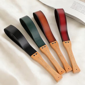 May include: Four leather paddles in black, green, brown, and red, each with a wooden handle. The handles have a light wood finish with decorative etched designs. The paddles are arranged on a white surface.