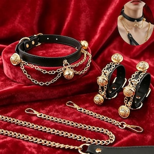 Pode incluir: Uma coleira de couro preta e dois punhos a condizer, cada um com correntes de metal dourado e berloques de sino em filigrana. O conjunto inclui correntes de elos dourados destacáveis com fechos lagosta para conectar as peças.