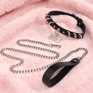 May include: A black collar with silver-toned metal rings and a bone-shaped tag engraved with the word Bitch, paired with a matching black leash featuring a silver-toned metal chain section and a black handle with a silver-toned metal ring.