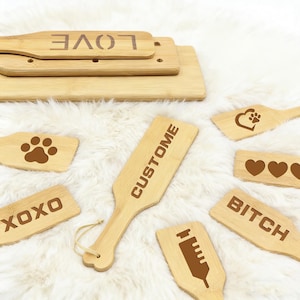 May include: A collection of bamboo paddles and small wooden boards with various engraved designs and text. The text includes "LOVE", "CUSTOM", "XOXO", and "BITCH". Other designs include a paw print, hearts, and a syringe.