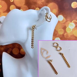 Puede incluir: Pendientes dorados exhibidos en una cabeza de maniquí blanca. Los pendientes incluyen pendientes de aro dobles, pendientes de cadena y un ear cuff dorado. Se muestran pendientes adicionales en una exhibición separada.