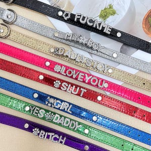 May include: A selection of brightly coloured pet collars with rhinestone detailing. The collars are in a range of colours, including black, silver, gold, pink, red, blue, green, and purple. Each collar has a different word or phrase spelled out in rhinestones, such as "FUCKME" and "Bitch".