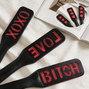 Puede incluir: Paletas de cuero negro con letras rojas. Una paleta dice "XOXO", otra "LOVE" y una tercera "BITCH". Una inserción más pequeña muestra paletas con diseños de estrellas y corazones.