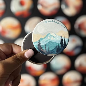 May include: A round magnet with a scenic illustration of a mountain range, trees, and a moon, with the word "Washington" printed on it. The magnet is held up, revealing its magnetic backing. Other magnets with similar designs are in the background.