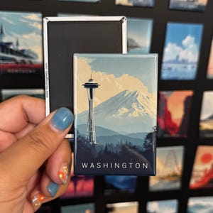 May include: A rectangular magnet featuring a scenic view of Washington State. The design includes the Space Needle, a mountain, and the word "WASHINGTON" in white. The magnet is held in front of a black background with other magnets.