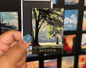 May include: A rectangular magnet featuring a landscape scene of Georgia with a large tree and Spanish moss. The word "GEORGIA" is printed at the bottom. The magnet is held against a black surface with other magnets.