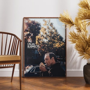 Op de afbeelding: Een ingelijste filmposter voor "The Usual Suspects" met een rokende man tegen een achtergrond van bomen en een auto. De poster zit in een houten lijst en leunt tegen een witte muur. Een houten stoel en vaas staan ook in de afbeelding.