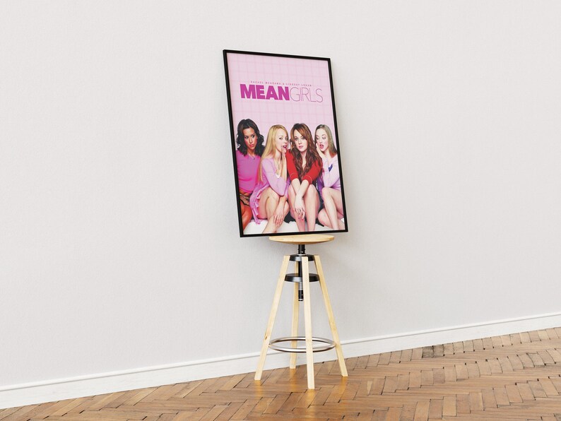 May include: A framed movie poster for the film "Mean Girls" featuring four young women in pink clothing. The poster is displayed on a wooden stool.